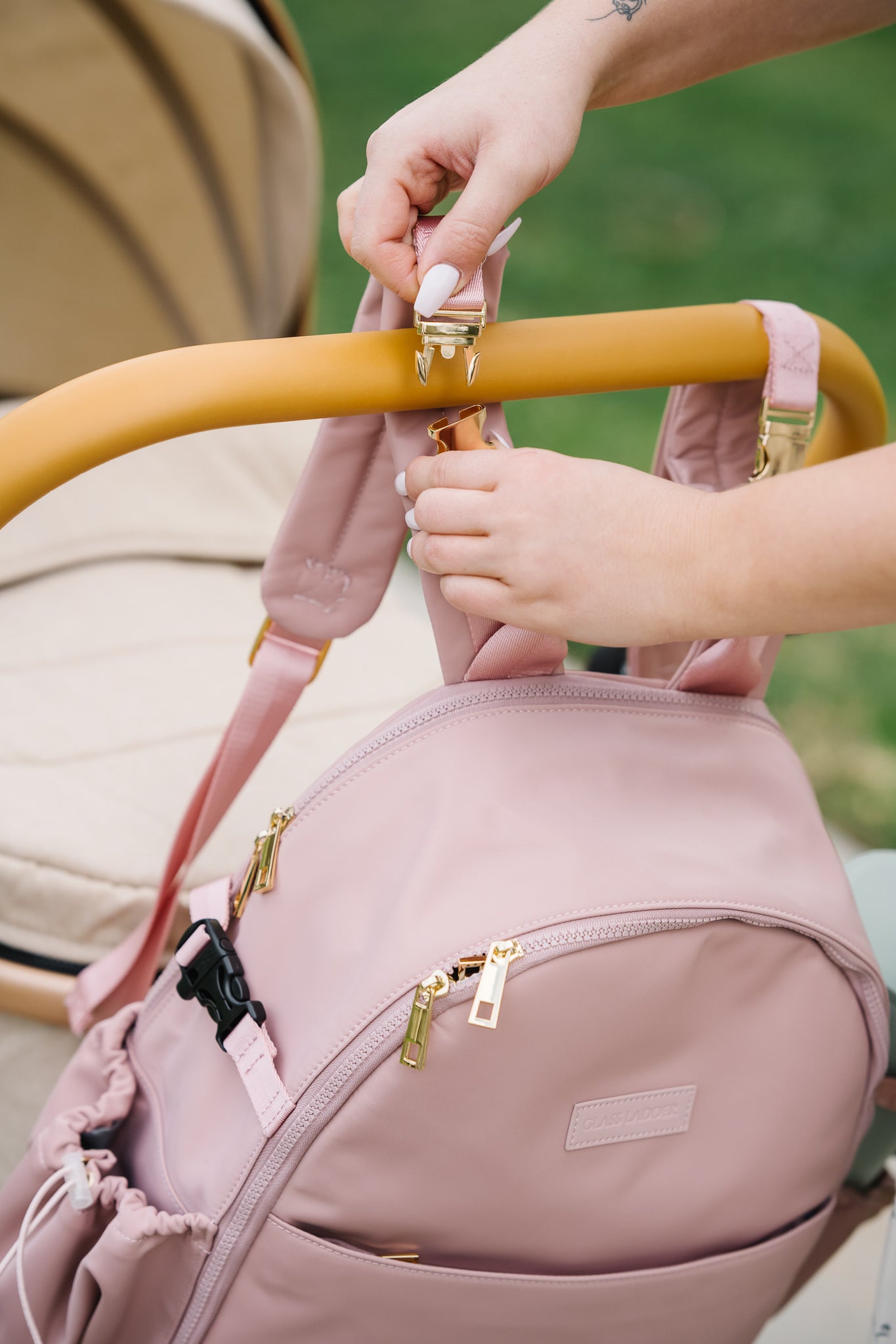 Someone attaches the Amanda Backpack by Glass Ladder, measuring 12 x 6 x 15, to a stroller with hooks. The stroller has a yellow handle and beige seat. The pink backpack with gold zippers and multiple pockets is shown against blurred greenery.
