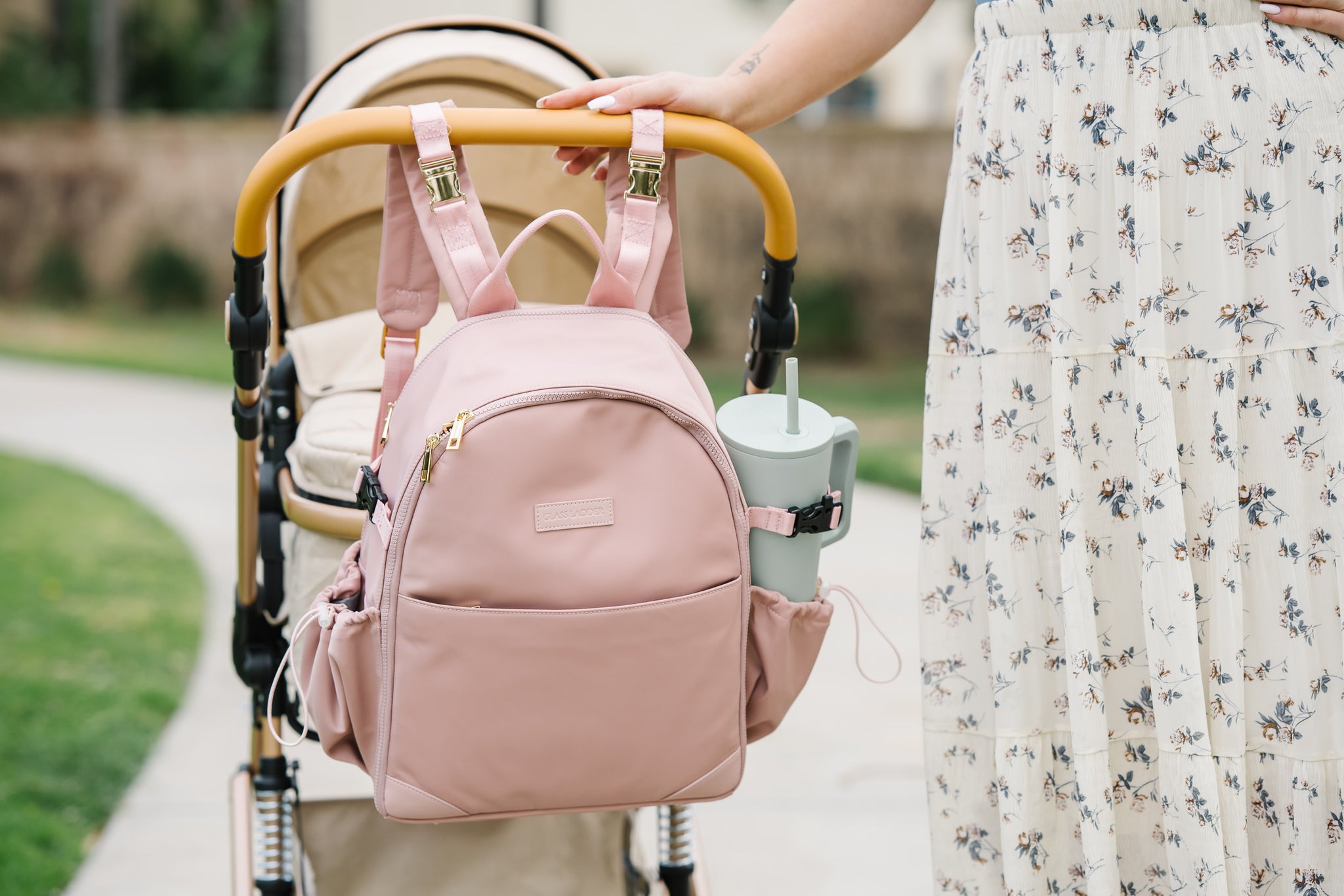 A person in a floral skirt holds a stroller with a tan shade, the Amanda Backpack — Pink Mimosa by Glass Ladder (L 12 x W 6 x H 15) hanging from the handle. A gray tumbler sits in the cup holder as they stroll on a paved path surrounded by vibrant green grass.