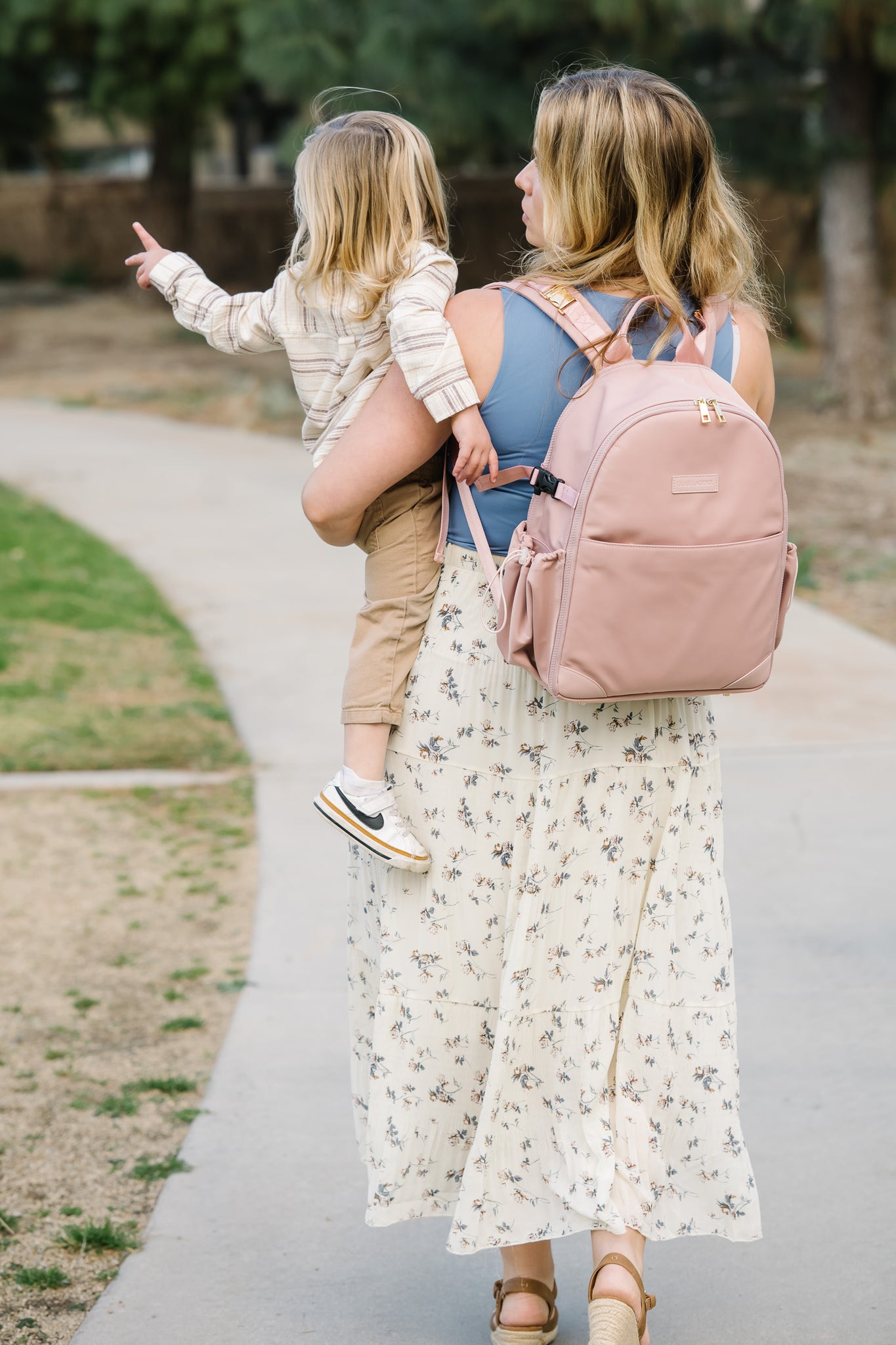 A woman in a floral dress carries a child with one arm, her Glass Ladder Amanda Backpack in Pink Mimosa (L 12 x W 6 x H 15) is snug on her shoulders, as they walk through a park path lined with trees. The child eagerly points ahead, their journey framed by lush grass and greenery.